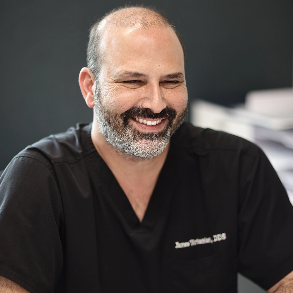Dr. James Vartanian, DDS, smiling warmly in his dental clinic, wearing a black dentist's uniform with his name embroidered on the chest.