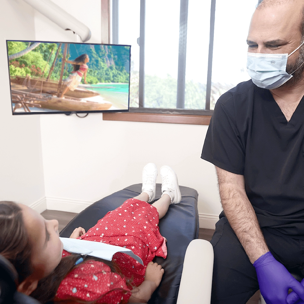 A young girl in a dental chair watches a TV screen showing a peaceful nature scene while Dr. James Vartanian, wearing a mask, looks on attentively, ensuring her comfort during the dental examination.