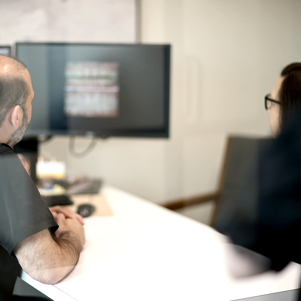 A male dentist sitting in front of a computer screen discussing dental images with a female patient or colleague, pointing towards the screen in a modern dental office setting.