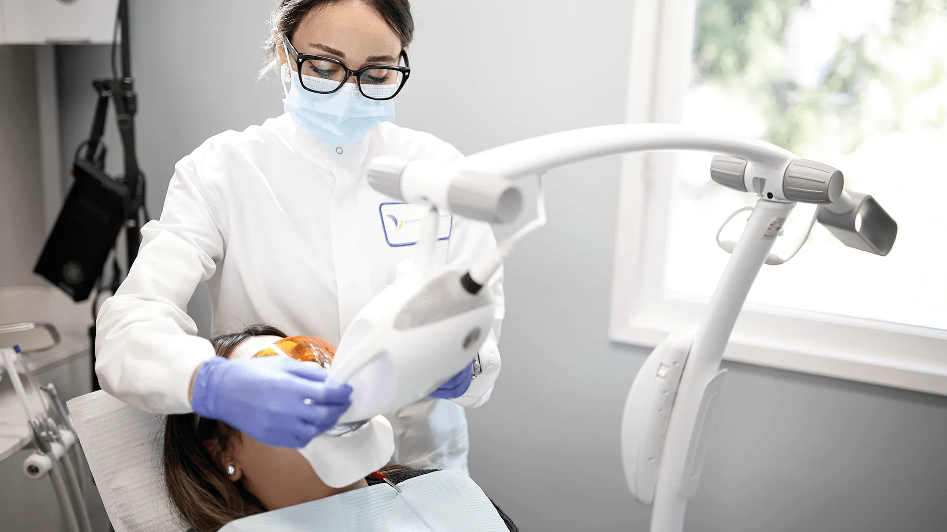 The female dentist is seen using an advanced LED light device to enhance the teeth whitening process. The patient lies comfortably in the dental chair with protective eyewear as the light activates the whitening gel.