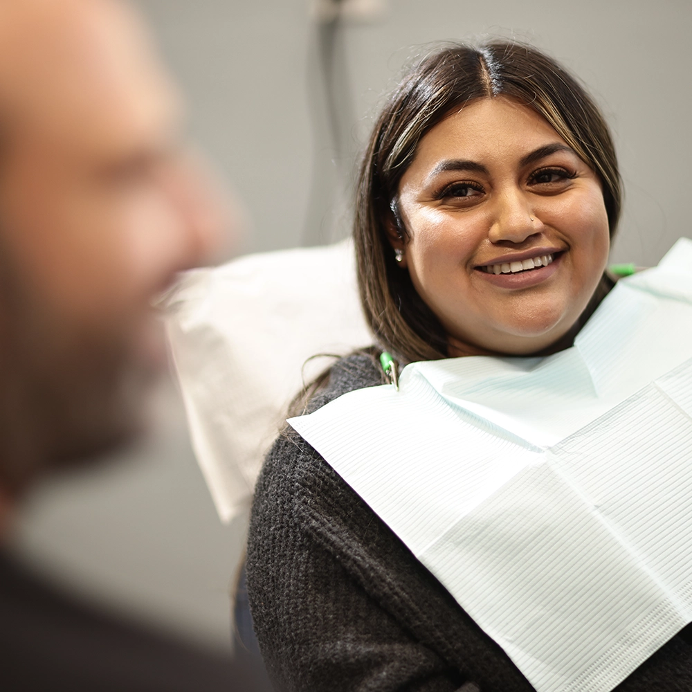 A male dentist with a bald head, viewed from behind, discusses dental images on a computer monitor with a female patient with long dark hair, who is partially visible. The scene occurs in a modern dental office setting.