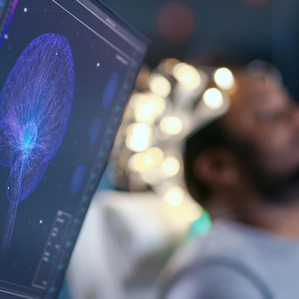 A middle-aged man in a dark blue shirt, sitting under a neurological examination device, observing a glowing blue neuron structure on a screen in a dimly lit room.