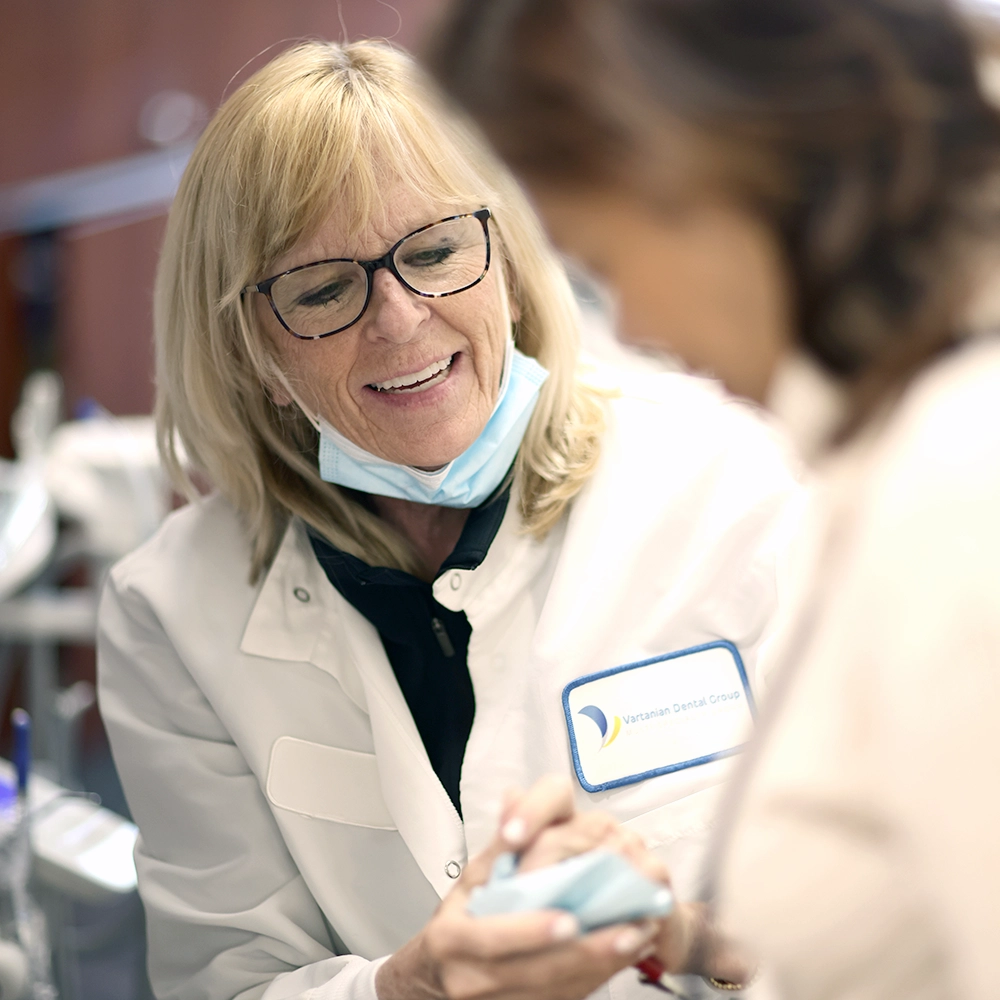 A female dentist with blonde hair, wearing glasses and a white lab coat with the Vartanian Dental Group logo, is smiling warmly while engaging with a patient. 