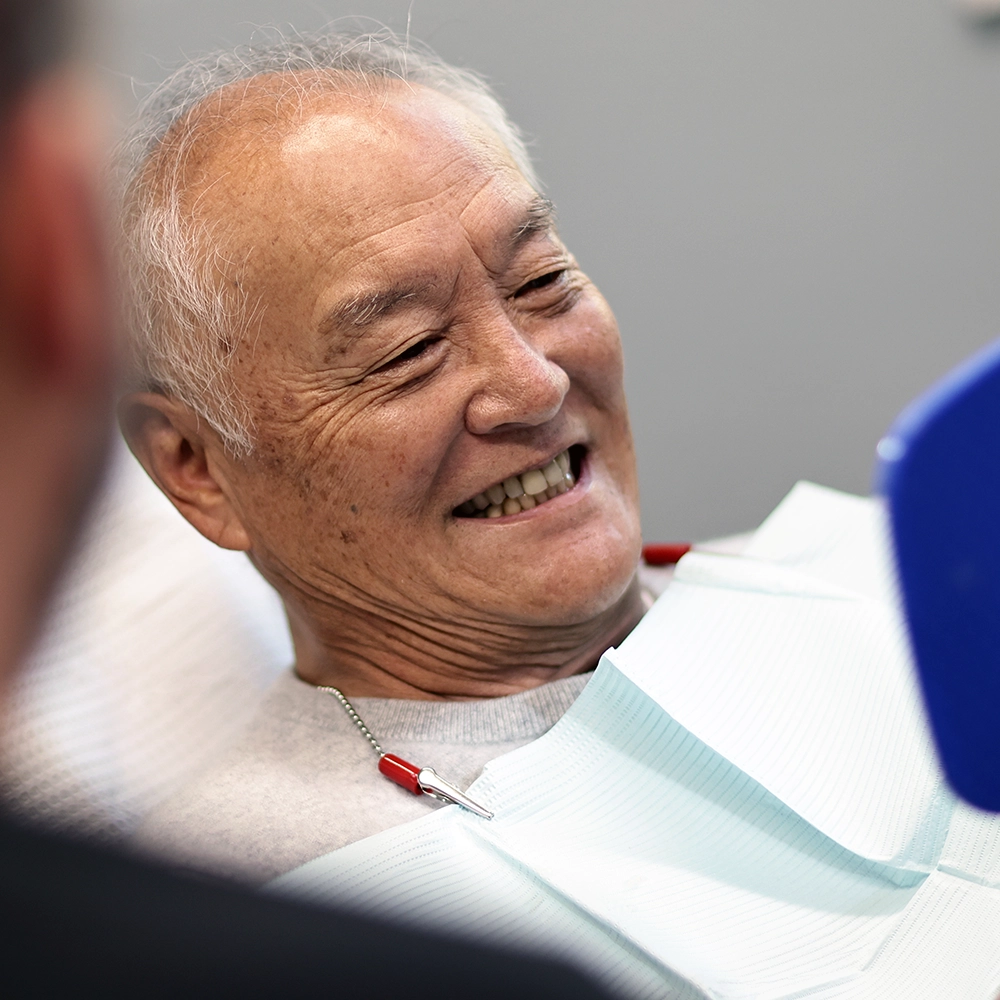 An elderly man laughs joyfully during a dental appointment, seated in a dental chair with a dentist in a dark shirt and glasses examining him. 