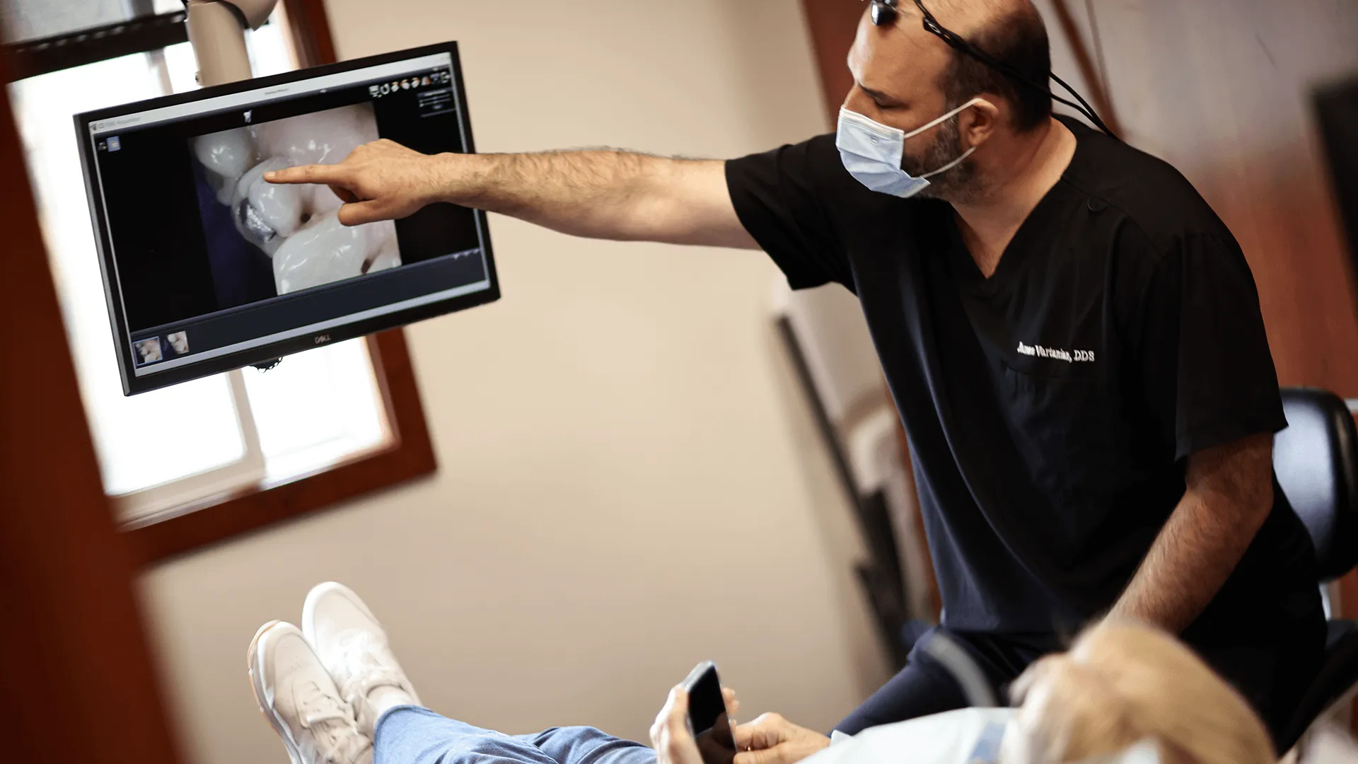 A male dentist in a black clinic uniform is explaining dental images on a monitor to a patient whose face is out of frame.