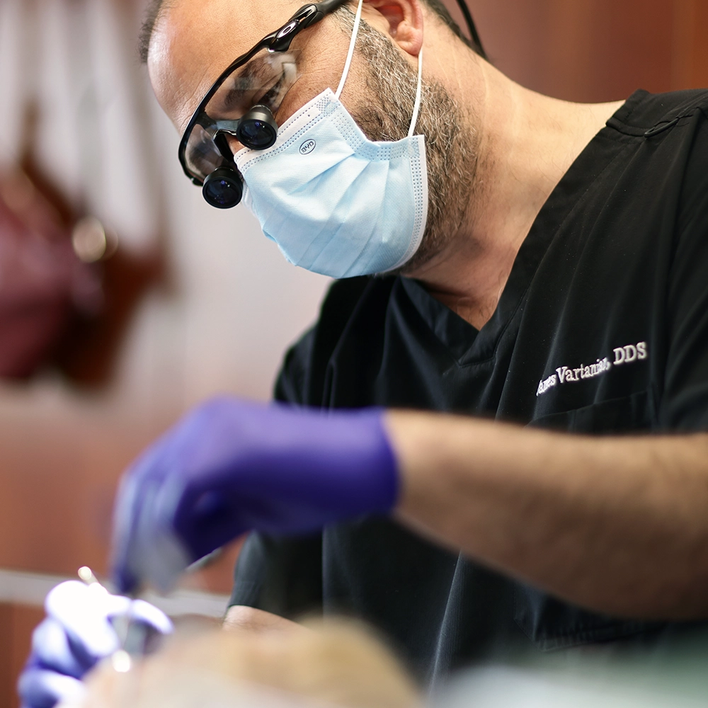 Dr. James Vartanian, wearing a protective mask and magnifying loupes, is focused on performing a dental procedure. He is dressed in a black dental scrub, and the image captures the precision and care taken during the treatment.