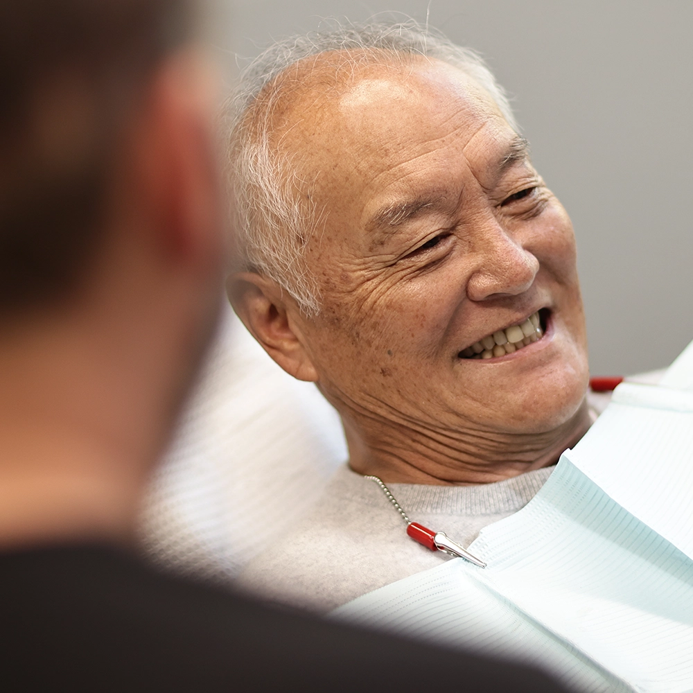  An elderly Asian male patient laughs heartily while seated in a dental chair, wearing a light gray bib. The dentist, seen from the back, engages with the patient, creating a relaxed and cheerful atmosphere.