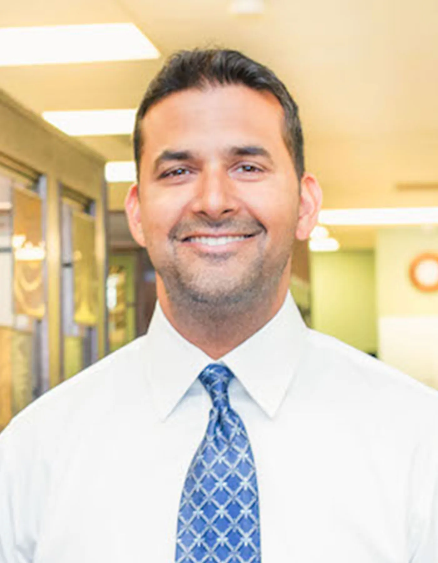 Portrait of Dr. Chopra, a dentist, smiling in his office. He wears a light blue shirt with a patterned blue tie, conveying a professional yet approachable demeanor.