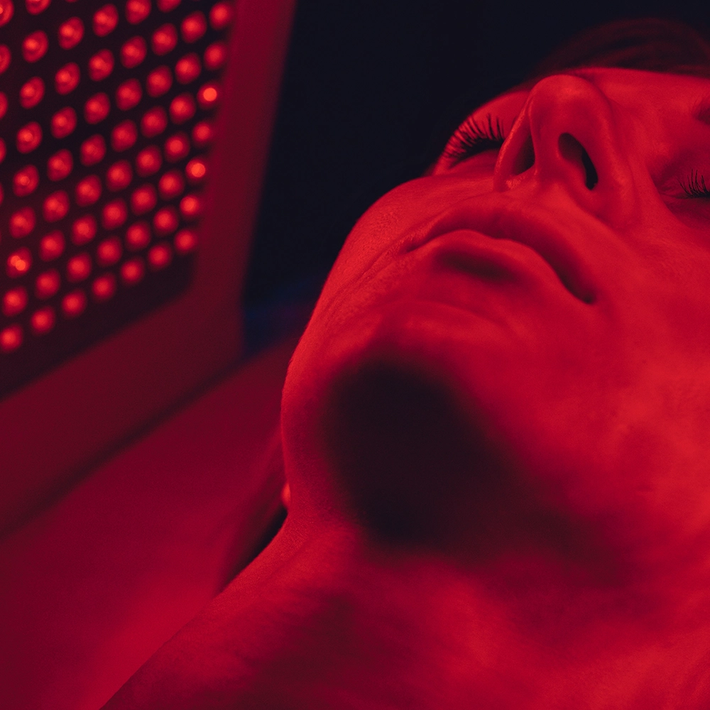 A woman lying down with her eyes closed, undergoing red light therapy, illuminating her face with a deep red glow, in a clinical setting.