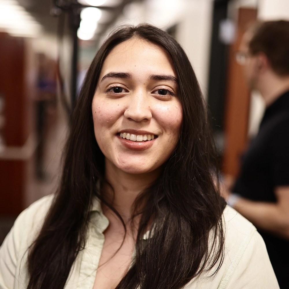 A close-up of a smiling young woman with long dark hair and a light green shirt, standing in a dental office. She appears cheerful and relaxed.