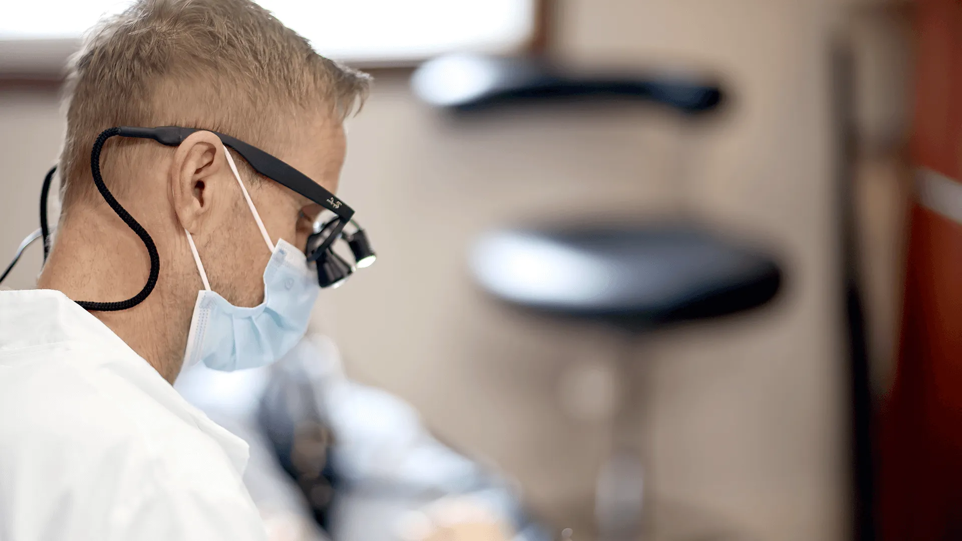 A male dentist wearing a surgical mask and magnifying glasses focused on examining a patient’s teeth, with dental equipment in the background.