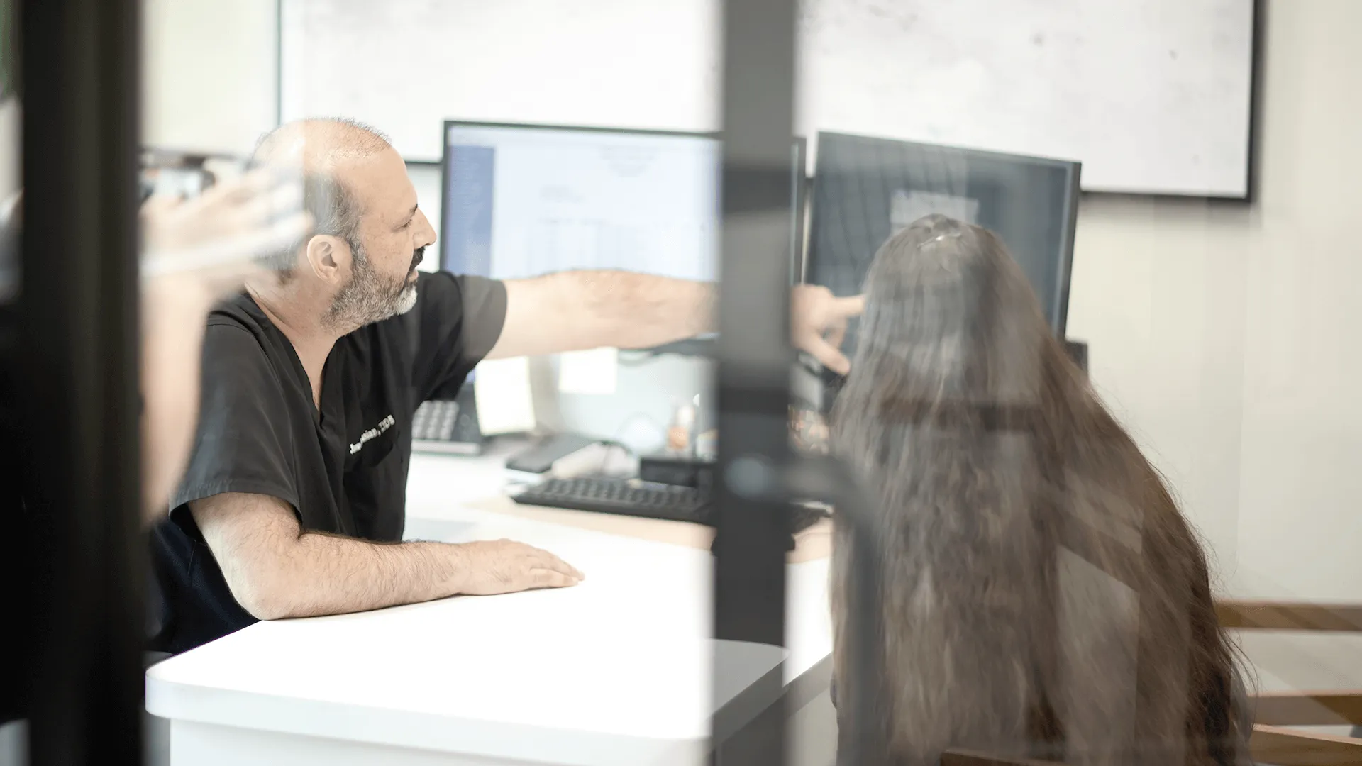 Dr. Vartanian in a black uniform on his shirt, points to a screen displaying patient information to a female colleague with long hair in a modern dental office.