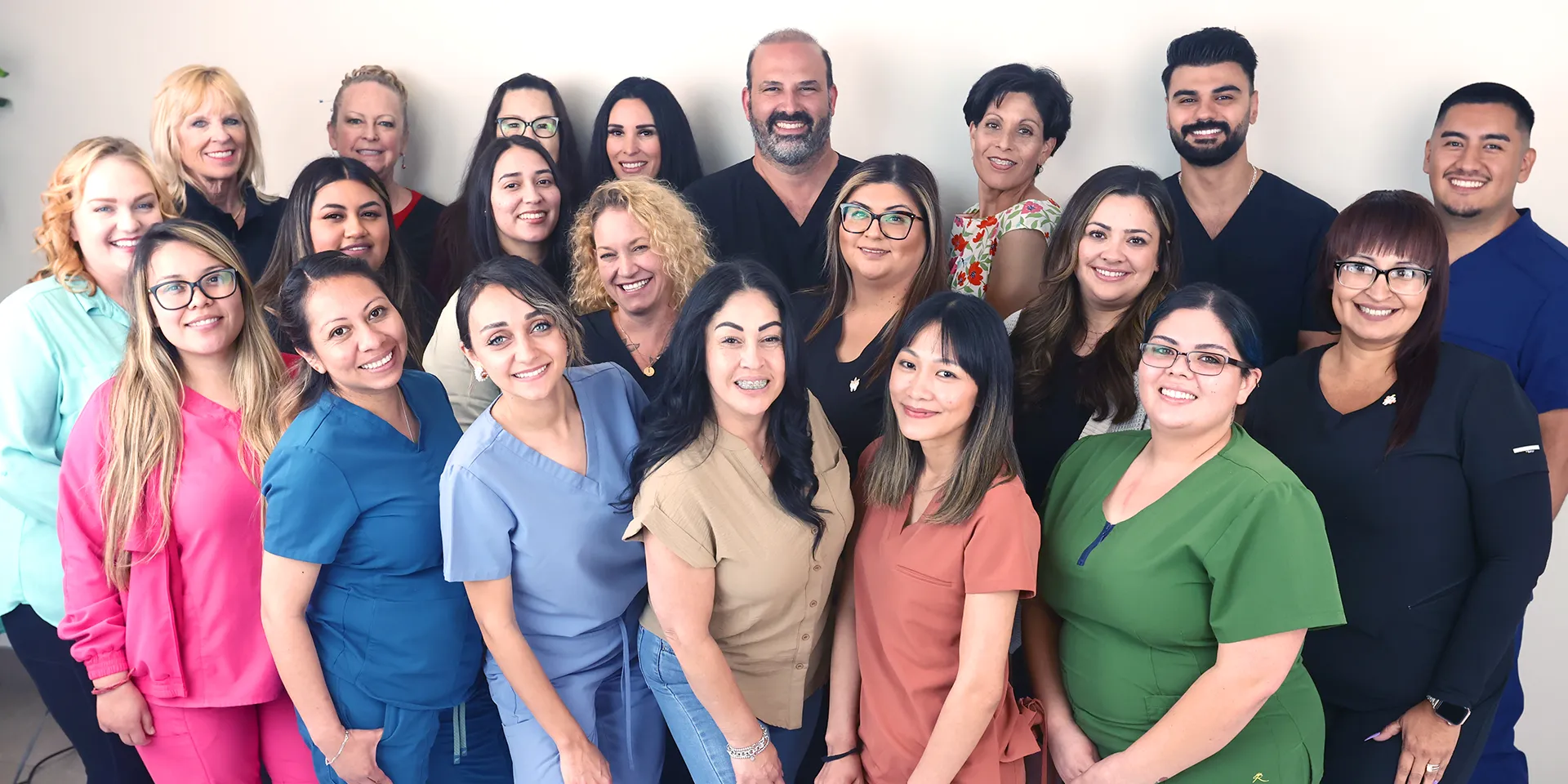 Group photo of Dr. James Vartanian and his diverse dental team, comprising of smiling members in various colorful medical scrubs. They are standing in two rows within the clinic, showcasing a mix of ethnicities and genders, radiating a friendly and professional atmosphere.