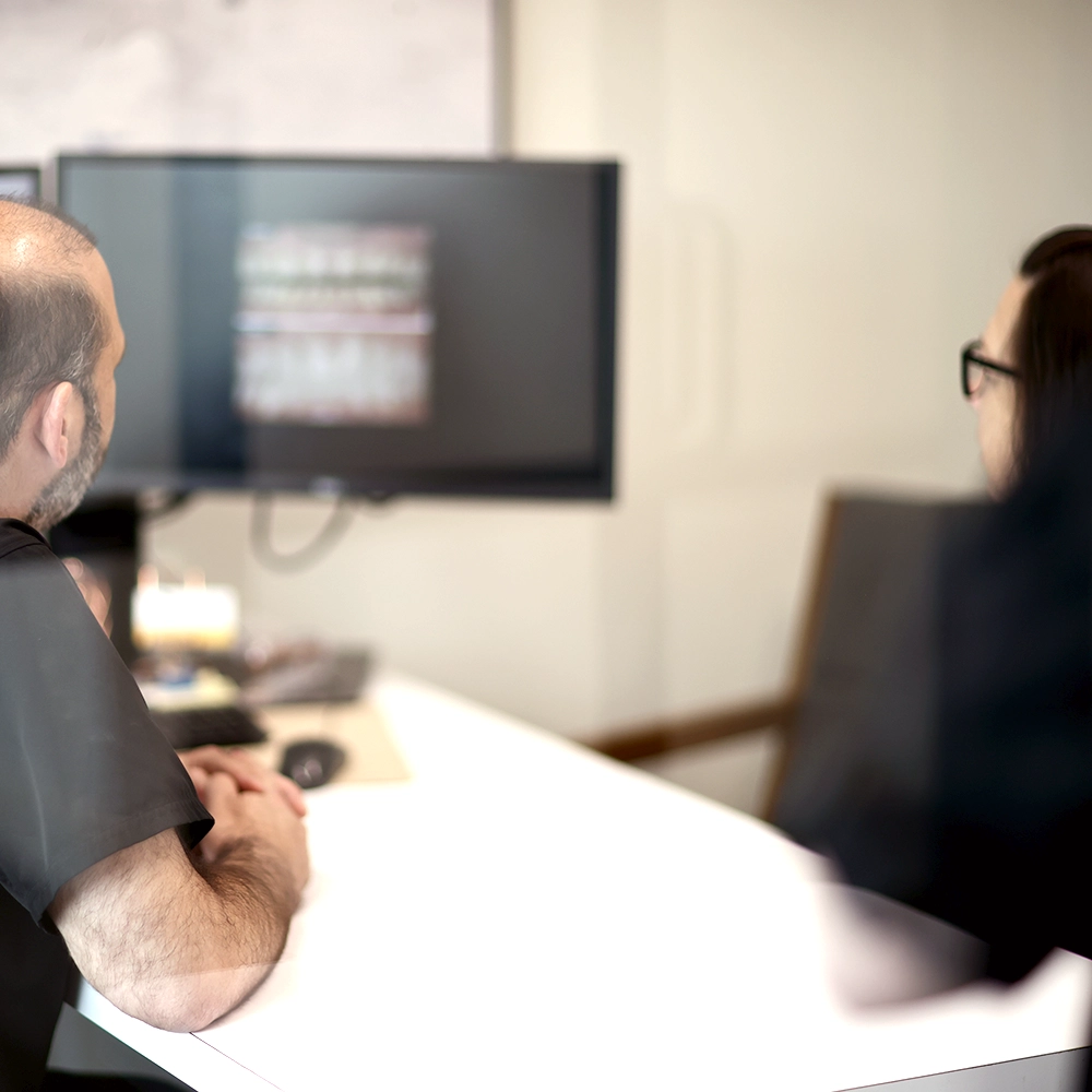 Scene inside a modern dental office, where a male dentist in black scrubs and a blue mask is engaged in a discussion with a patient or colleague, pointing at a dental X-ray displayed on a monitor, highlighting details of the dental structure.