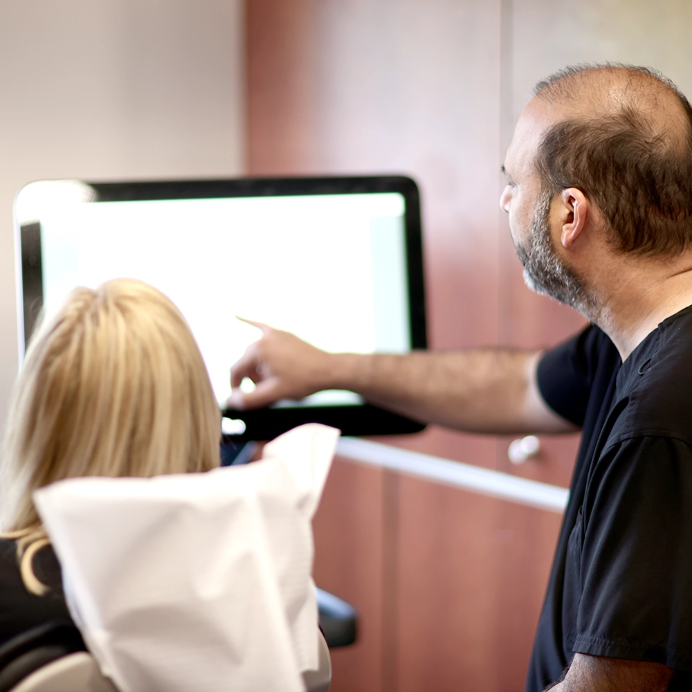 A male dentist with a beard, reviewing a large white screen pointing to specific dental images, explaining the procedure to a female patient wearing a dental bib.