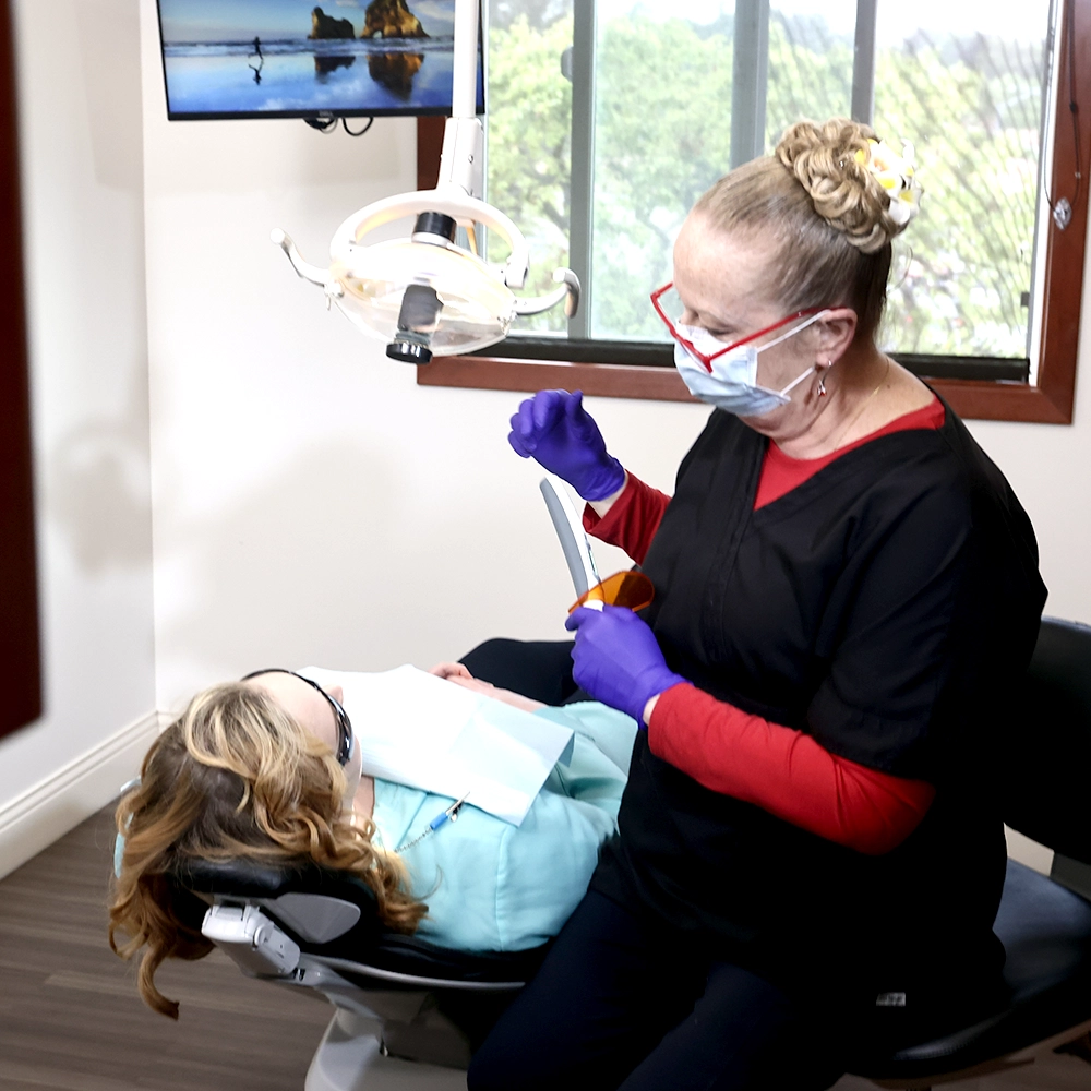 Dental treatment in progress by a senior dental professional in black and red attire, working on a patient reclined in a dental chair. The room has a relaxing ambiance with a scenic view through a window, ensuring a comfortable patient experience.