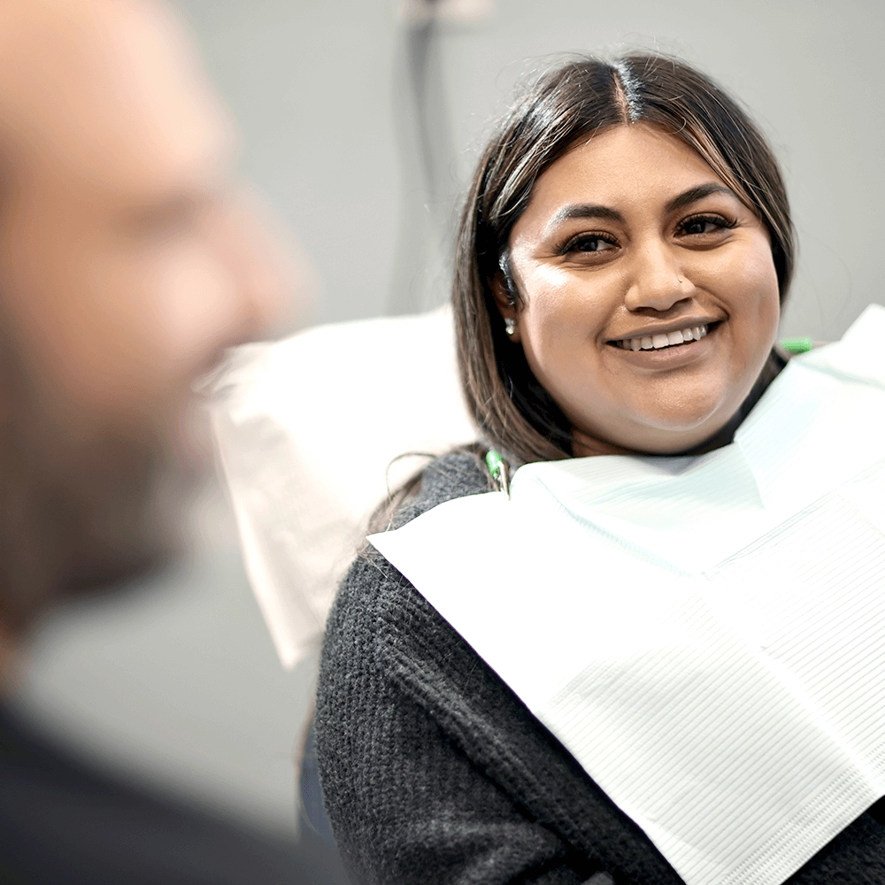 Dr. James Vartanian, smiling and interacting with a female patient wearing a grey sweater, seated in the dental chair. She appears relaxed and happy, protected by a dental bib, as Dr. Vartanian conducts a consultation.