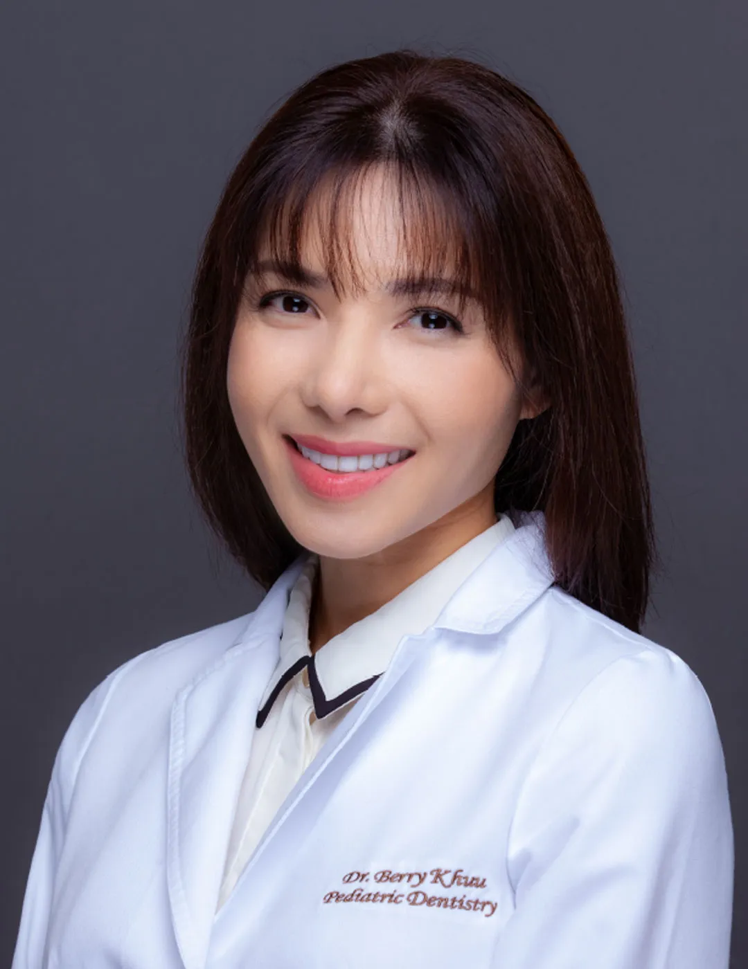 A professional portrait of Dr. Berry Khan, an Asian female pediatric dentist, wearing a white coat embroidered with her name and specialty