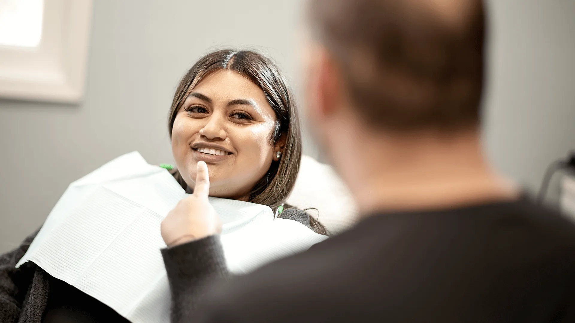 A cheerful woman in a dental chair wearing a bib is smiling at her dentist during a consultation. The dentist, a male with a beard and wearing a surgical mask and gloves, is out of focus in the foreground.